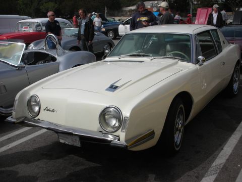 1998 Ford Crown Victoria - A cream 1963 Studebaker Avanti is parked at a car meet, with other vehicles visible in the background.