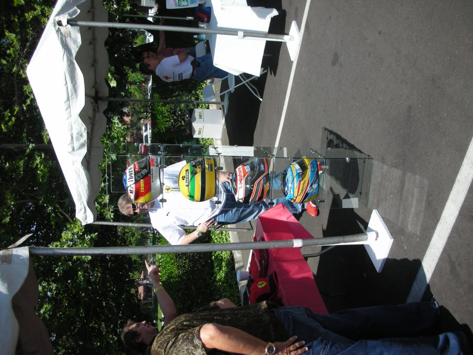 Several racing helmets are displayed on a rack under a tent. People are standing around the display