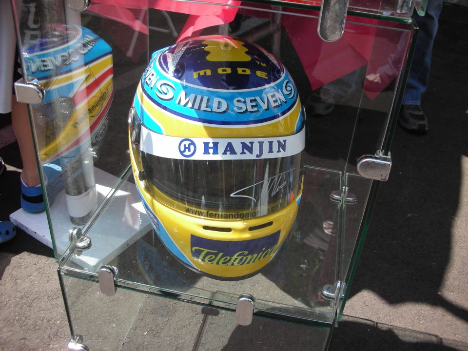 Yellow and blue racing helmet with "Mild Seven" and "Telefonica" text, displayed in a glass case