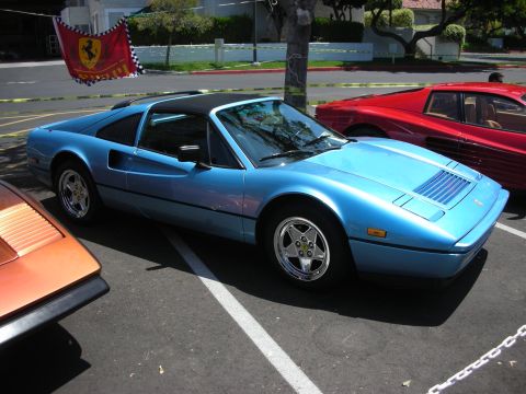 1998 Ford Crown Victoria - A blue Ferrari 308 GTS is parked in a lot next to other cars, with a Ferrari flag overhead.