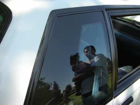 1998 Ford Crown Victoria - Man and child reflected in the window of a car parked outdoors on a sunny day.