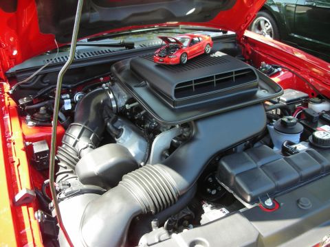 1998 Ford Crown Victoria - A red toy Mustang sits atop the engine of a red car, with another green car visible in the background.