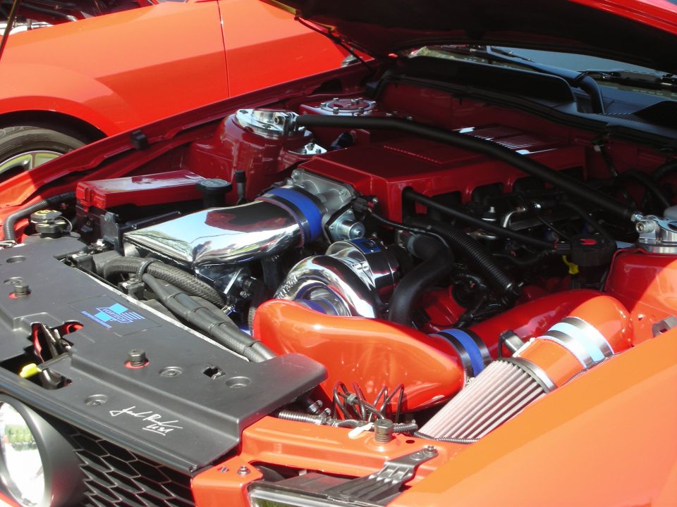 engine bay with turbocharger, chrome intake, and red engine covers. Another red car is visible in the background
