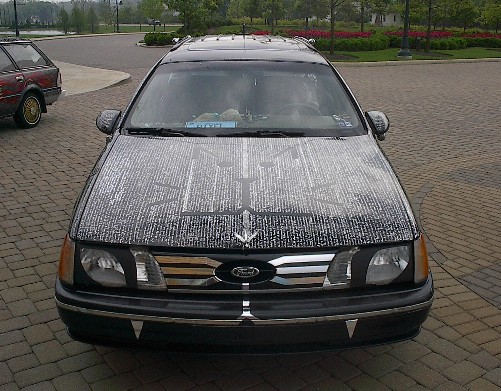 Black Ford Taurus sedan front view with hood bra and headlight covers, wet hood