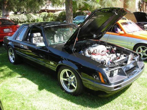 1998 Ford Crown Victoria - Black 1987 Ford Mustang 5.0 on display with hood open, parked on grass.