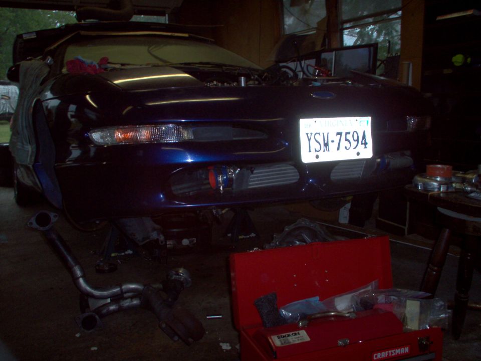 Blue coupe on jack stands with hood open, front end visible. Intercoolers are mounted behind the bumper. Tools and parts are scattered around