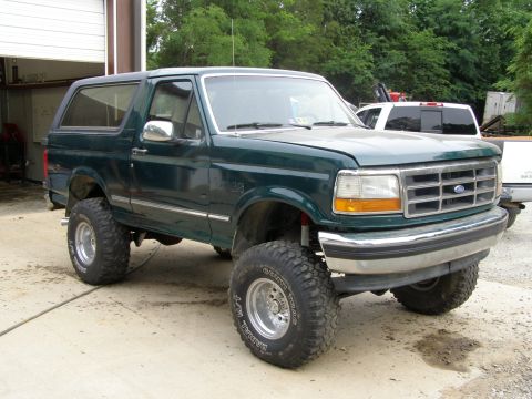 1993 Ford Bronco - A lifted green Ford Bronco sits outside a garage, showing off its off-road tires and suspension modifications.