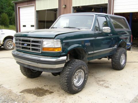 1993 Ford Bronco - A lifted green Ford Bronco is parked outside a garage, with another white truck visible to the left.
