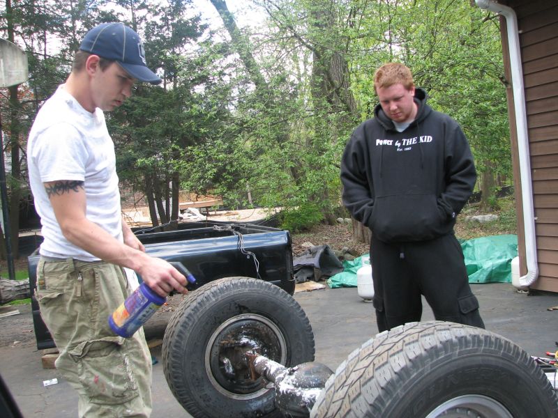 Two men are working on a truck bed and axle assembly outdoors. One man sprays a fluid onto the axle