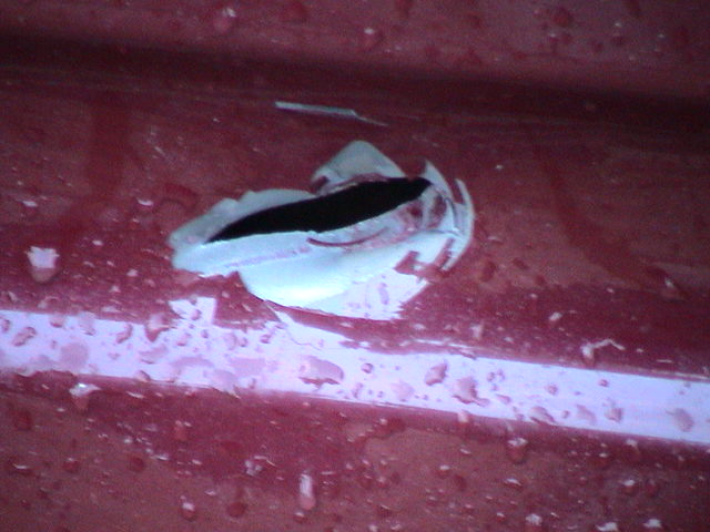 Close-up of puncture damage and chipped white primer on a dark red vehicle body panel with water droplets