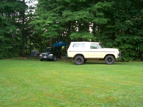1978 Ford Bronco - Two vehicles are parked on a grassy area near trees, with camping tents set up in the background.