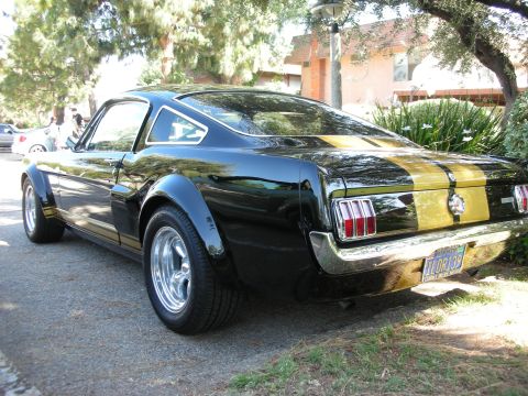 1998 Ford Crown Victoria - Rear view of a black 1965 Ford Mustang with gold racing stripes and aftermarket wheels.