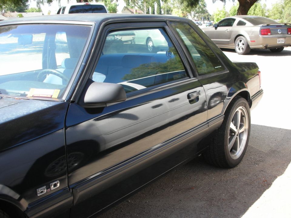 Black Ford Mustang with 5.0 badge on the fender, parked on asphalt. Another car is visible in the background