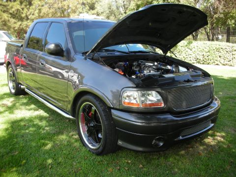 1998 Ford Crown Victoria - A modified Ford F-150 SVT Lightning is parked on grass with its hood open, revealing the engine bay.