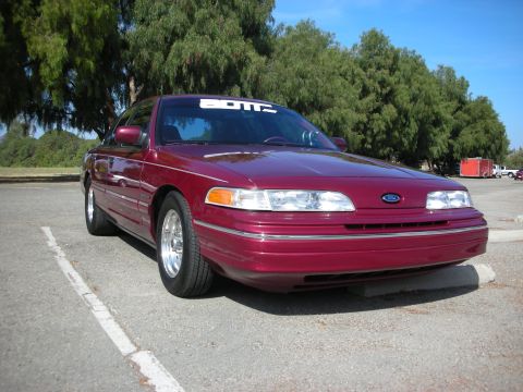 1998 Ford Crown Victoria - A maroon 1998 Ford Crown Victoria is parked in a lot, a white banner across the top of its windshield.