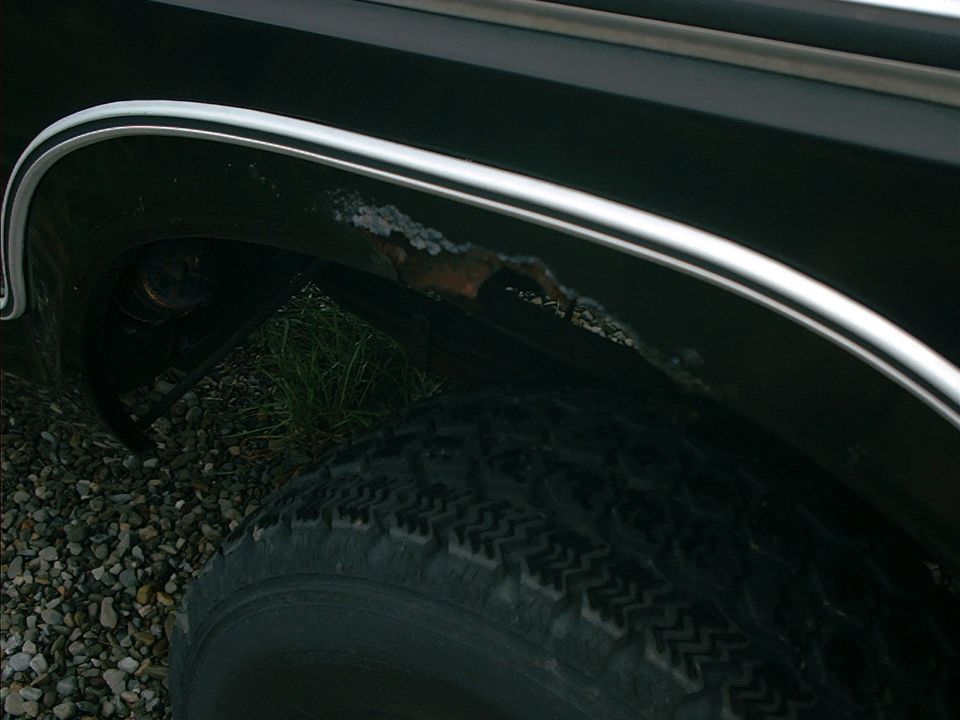 Rust holes and bubbling along chrome wheel arch trim on a dark SUV fender above the tire