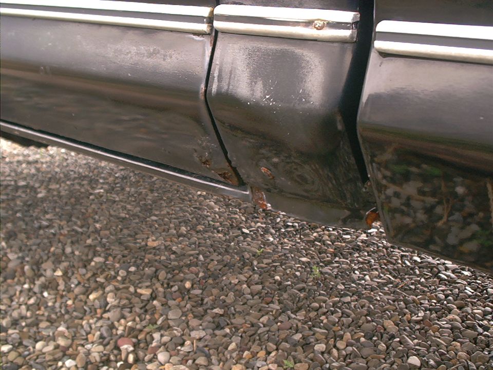 Close-up of rust spots on lower door edges and rocker panel of a black vehicle over gravel