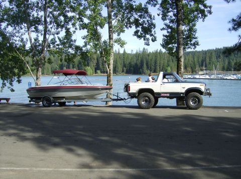 1986 Ford Bronco - A lifted white Ford Bronco tows a boat trailer near a lake with a marina in the background.