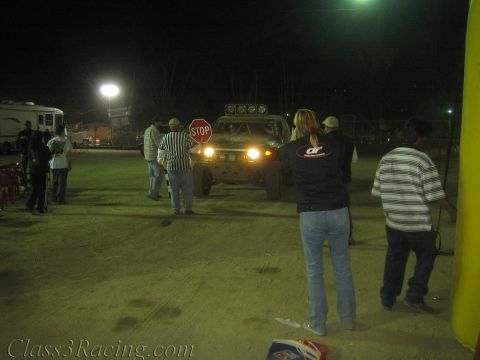 1990 Ford Bronco - off-road race vehicle with lit headlights pauses at a STOP sign at a night dirt staging area as a flagger and spectators stand nearby.