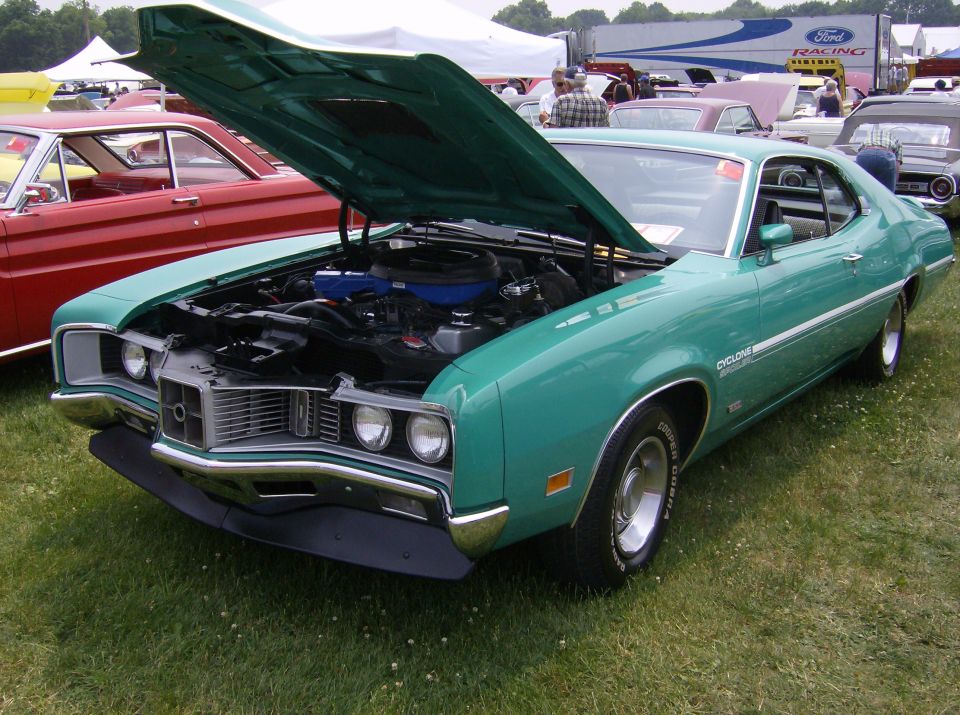 Green 1970 Mercury Cyclone with hood open, parked on grass at a car show. Another red car is to its left
