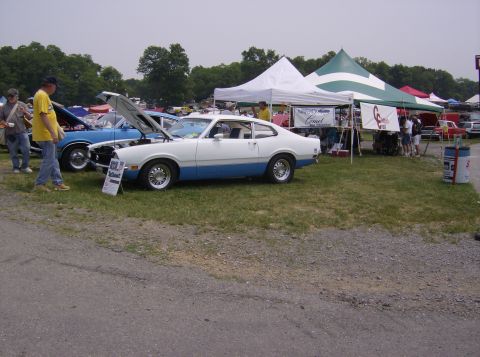 1970 Ford Ranchero - A white and blue Ford Maverick is displayed at an outdoor car show with tents and other vehicles in the background.