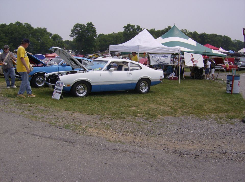 White and blue Ford Maverick with hood open, parked on grass at an outdoor event. Other cars and tents are visible
