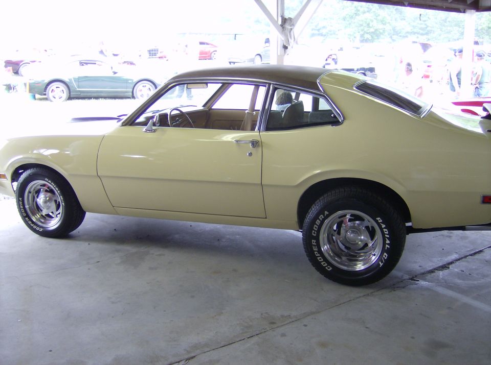 Yellow Ford Maverick coupe with a brown roof, seen from the side. Other cars are visible in the background