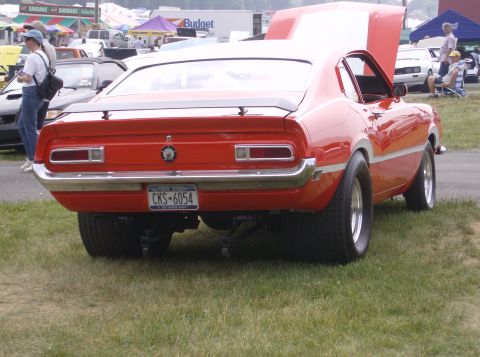 1970 Ford Ranchero - A red 1970 Ford Ranchero with a spoiler and wide rear tires is parked on grass, with other vehicles and tents in the background.