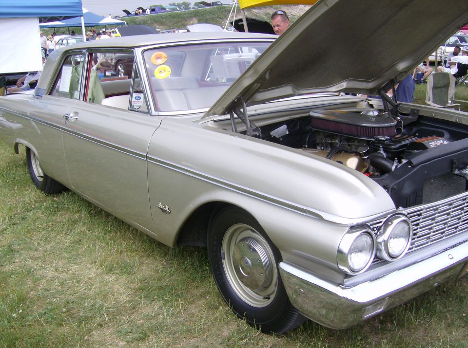 Silver 1960 Ford Fairlane with hood open, showing the engine. Parked on grass with tents and people in the background