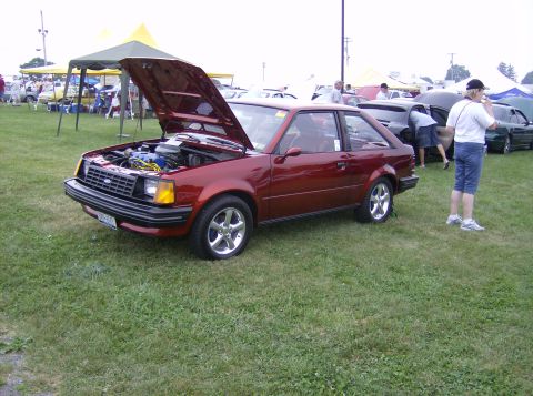 1970 Ford Ranchero - A red hatchback with its hood open is displayed on a grassy field, with other vehicles and tents in the background.