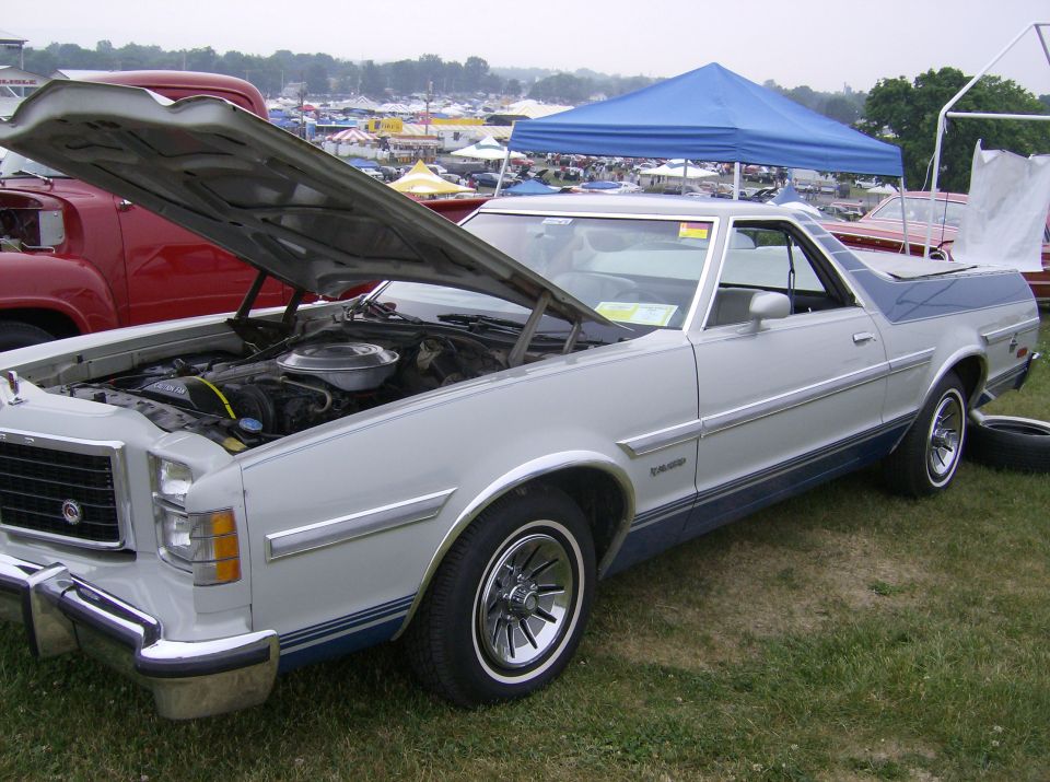 1977 Ford Ranchero, light gray with blue trim, hood open showing engine. Other vehicles and tents in background
