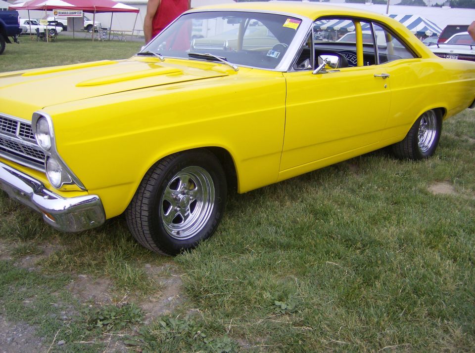 Yellow coupe with chrome wheels parked on grass. It has a prominent grille and dual headlights