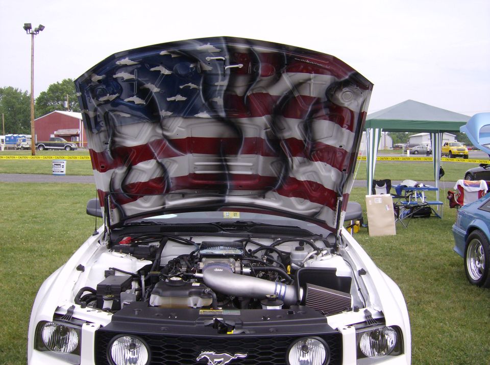 White car with hood open showing engine and American flag paint job. Other cars and tents in background