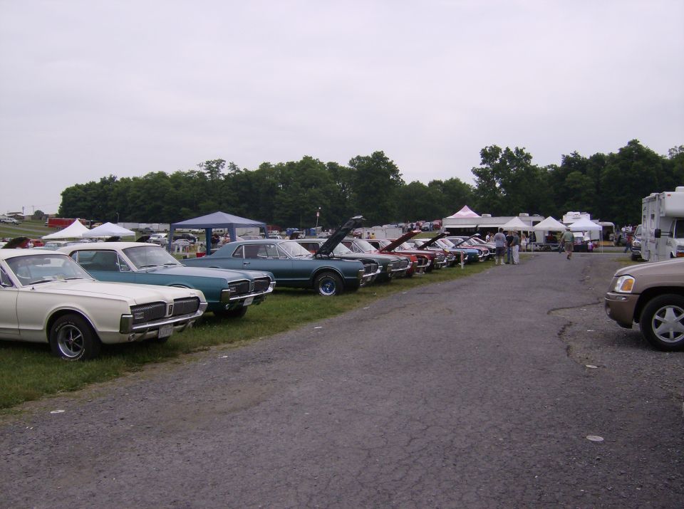 Several classic cars parked in a row at an outdoor event with tents and RVs in the background