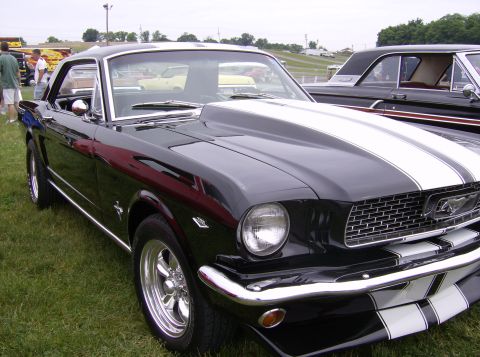 1970 Ford Ranchero - A black 1965 Ford Mustang with white racing stripes is parked on grass next to another classic car.