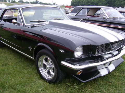 1970 Ford Ranchero - A black 1965 Ford Mustang with white racing stripes and a hood scoop is parked on grass, with another classic car behind it.