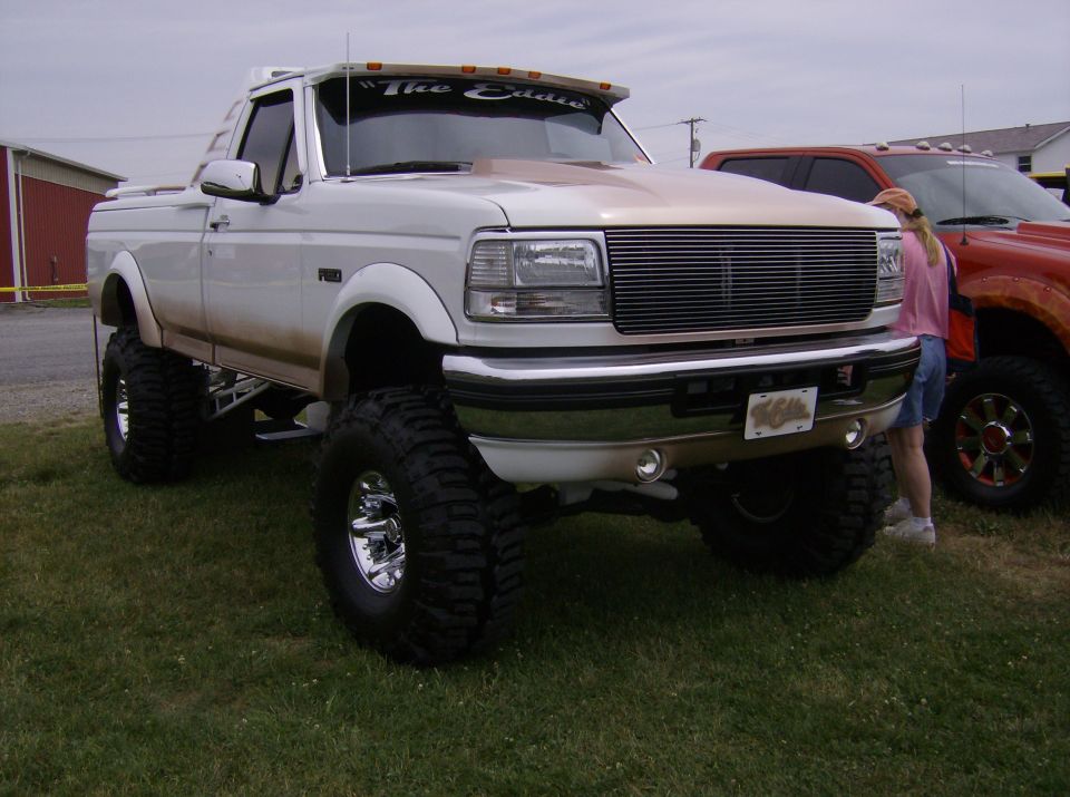 White lifted pickup truck with chrome grille and large tires, parked on grass next to an orange truck
