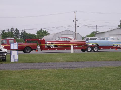 1970 Ford Ranchero - A red pickup truck transports two vintage cars on a trailer, with buildings in the background.