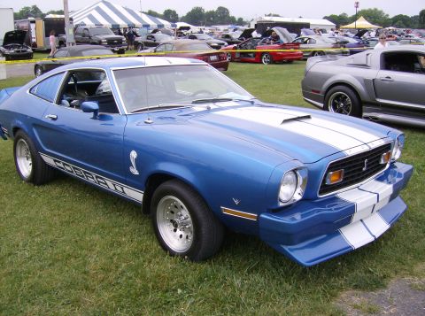 1970 Ford Ranchero - A blue 1976 Ford Mustang Cobra II is displayed at an outdoor car show with other vehicles in the background.