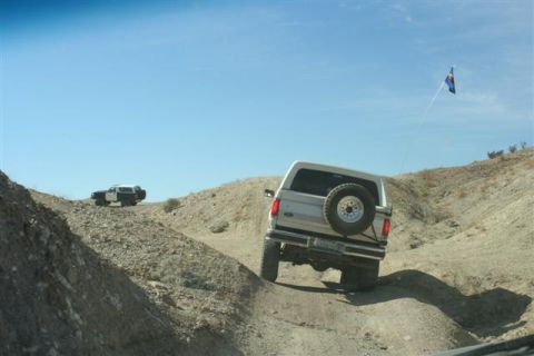 1994 Ford Bronco - Two SUVs navigate a dusty off-road trail under a clear blue sky, with a Colorado flag flying in the distance.