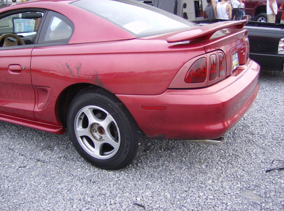 Red coupe with a spoiler and racing stripes on the rear quarter panel, parked on gravel