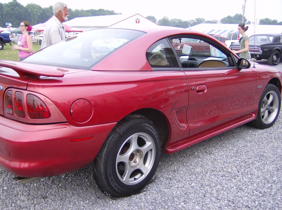 Red coupe parked on gravel, people in background