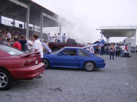 1970 Ford Ranchero - Cars are parked on gravel with a crowd watching an event under a cloudy sky.