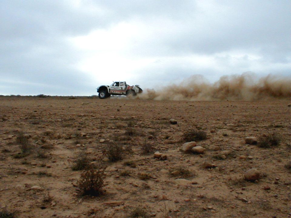 Off-road race truck driving on a dirt track, kicking up a large cloud of dust under an overcast sky