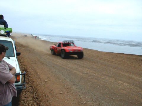 1996 Ford Bronco - A red off-road truck races along a dusty track beside the sea, with other vehicles and people in the distance.