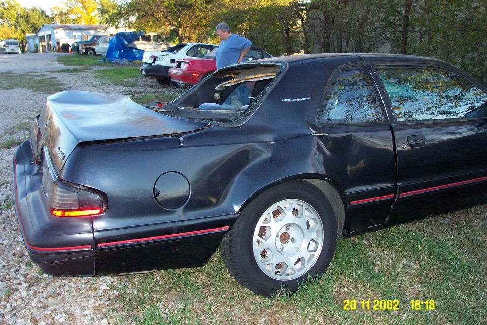 Rear 3q view of dark coupe with red stripe and white alloy wheels, rear glass missing, on gravel lot