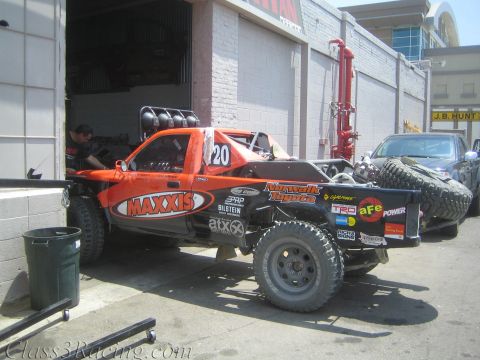 1990 Ford Bronco - orange and white off-road race truck covered in Maxxis and Toyota sponsor decals, numbered 20, sits outside an industrial shop bay.