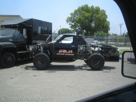 1990 Ford Bronco - black tube-chassis off-road race truck lettered 'Baja Motor Sports' sits in a lot beside a black box truck, viewed through a vehicle window.
