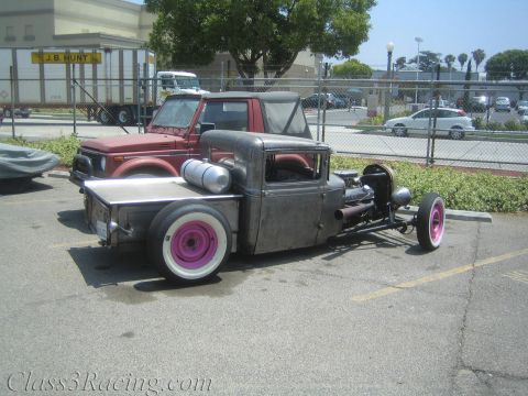 1990 Ford Bronco - bare-metal rat-rod pickup with chopped cab and pink-painted whitewall wheels sits in a parking lot, a red SUV behind it.
