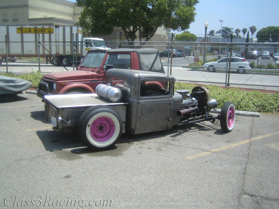 Bare-metal rat-rod pickup with pink whitewall wheels in parking lot